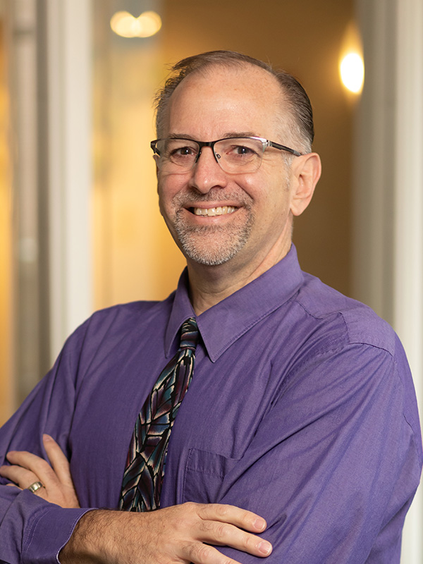 White presenting man with graying dark hair, glasses, goatee. purple shirt, multicolored tie.