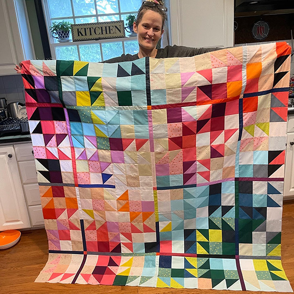 Blonde woman (Jill) holding a large multi-colored quilt. Quilt has many colors - triangle patterns stitched together.