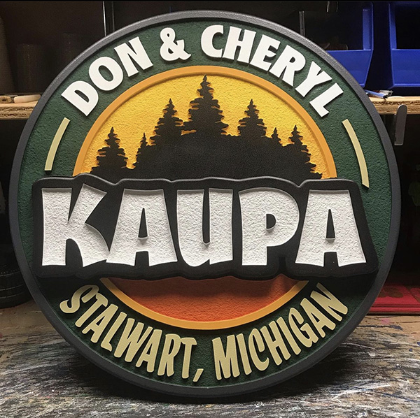Round sign by Mastyl Signs - white, green, black, yellow, and orange. "Don & Cheryl" around the top, "Kaupa" across the middle, and "Stalwart, Michigan" around the bottom. In the background are pine trees.