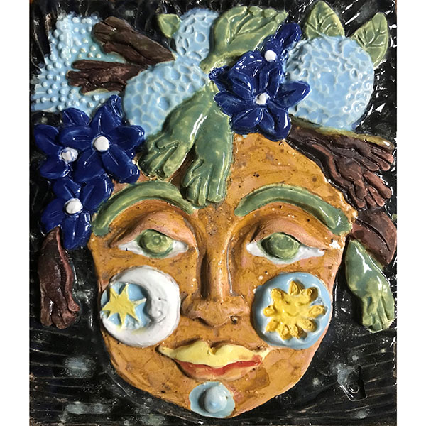 closeup of ceramic tile, showing a woman's face, with moon and sun symbology. the face is yellow, there are blue flowers and green leaves in the background. There are some hands in different shapes as well.